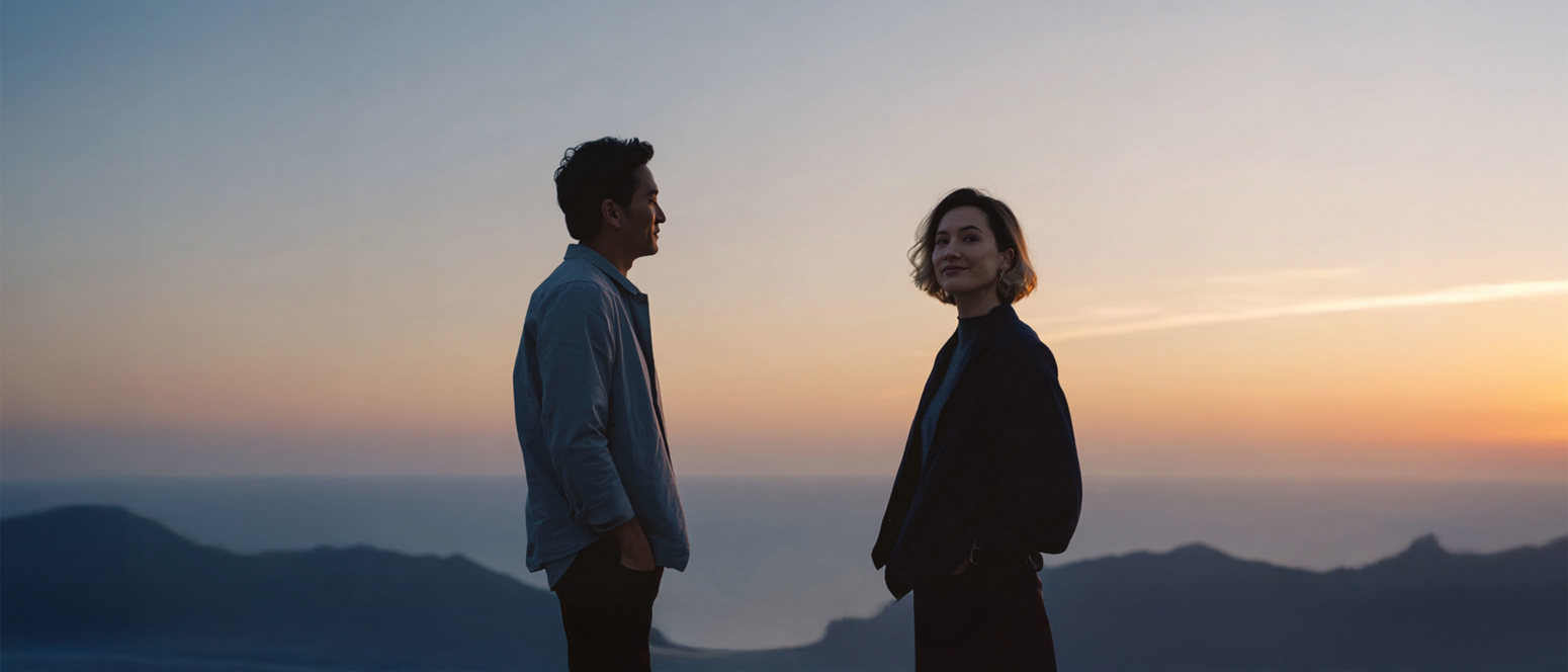 To mennesker står udendørs ved solnedgang med bjerge og hav i baggrunden.