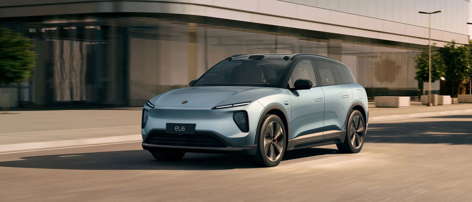 En lyseblå elektrisk SUV kører på en gade i byen foran en moderne glasbygning.